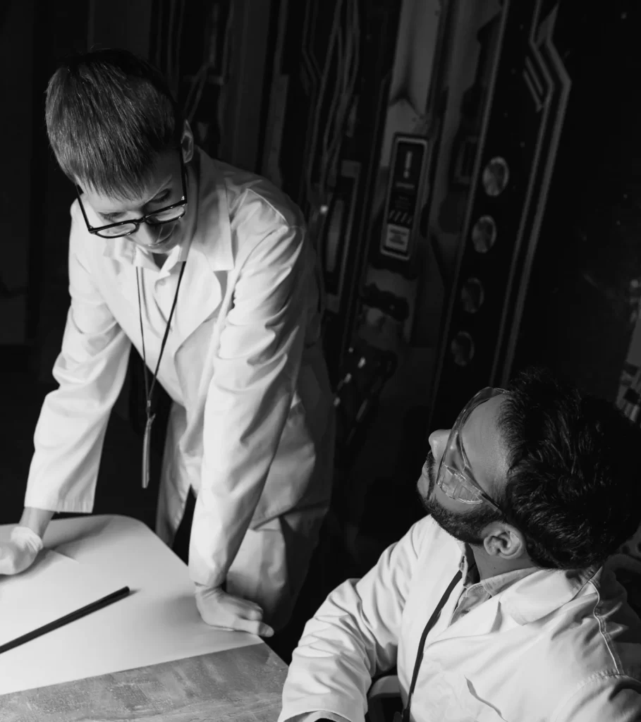 Two engineers in lab coats and safety glasses discuss a project at a table, reflecting collaboration in semiconductor technology development.