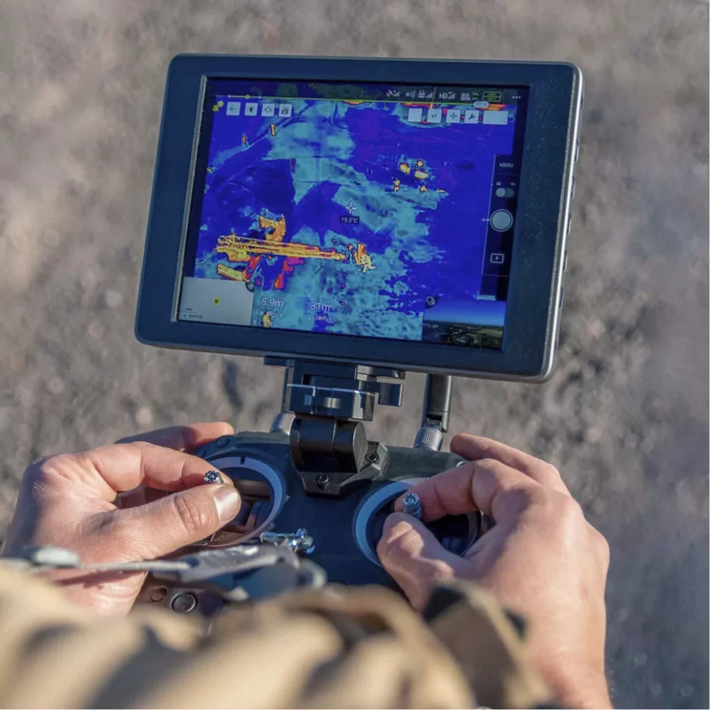 Hands operating a remote control with a screen displaying thermal imaging, relevant to aerospace and defense technology.
