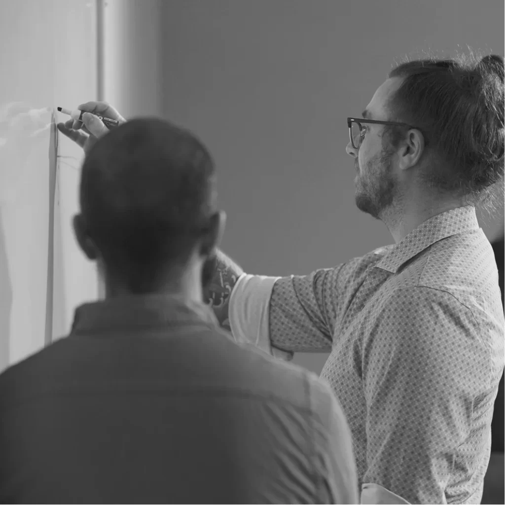 Two individuals collaborating at a whiteboard, one writing while the other observes, in a professional setting.