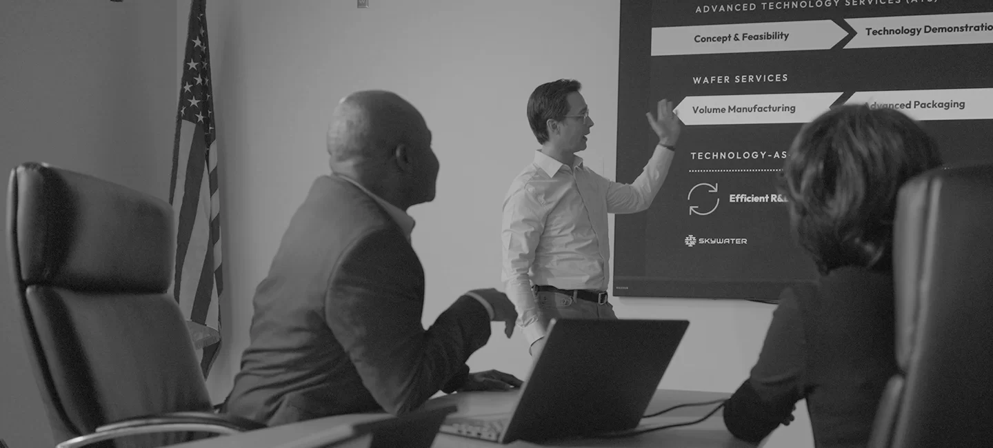 Three professionals in a conference room, with one person standing and gesturing towards a presentation screen displaying technology service phases.