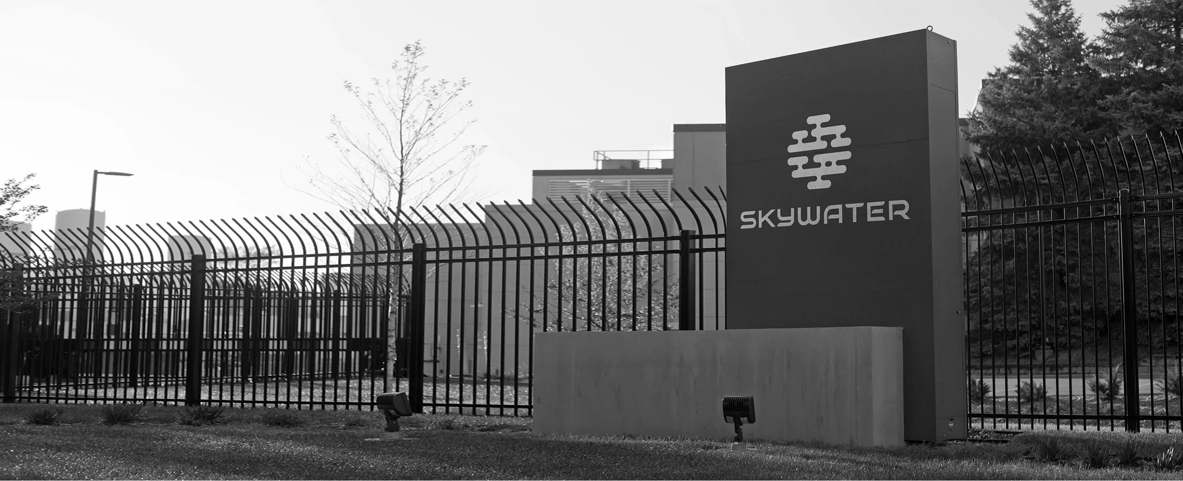 SkyWater Technology sign and building behind a fence, with trees and a clear sky in the background.