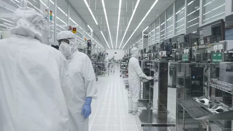 Technicians in cleanroom suits work in a semiconductor fabrication facility with advanced equipment lining the walls.