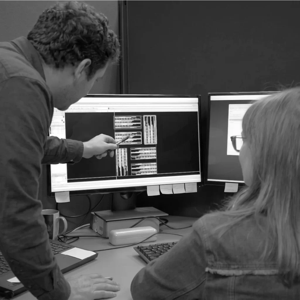 Engineer pointing at computer screens displaying semiconductor designs, collaborating with a colleague in an office setting.