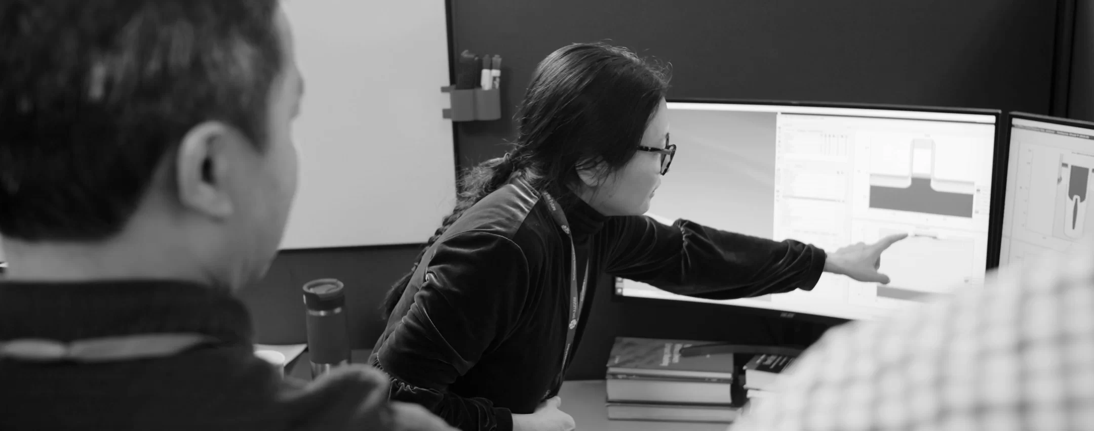 Engineer pointing at a computer screen displaying technical diagrams, collaborating with a colleague in an office setting.