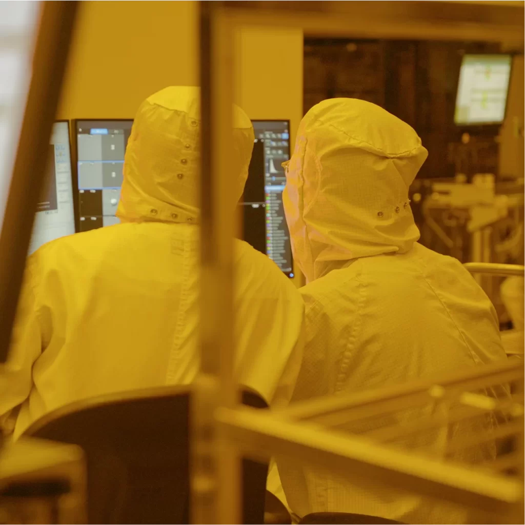 Technicians in cleanroom suits work at computer monitors in a semiconductor fabrication facility.
