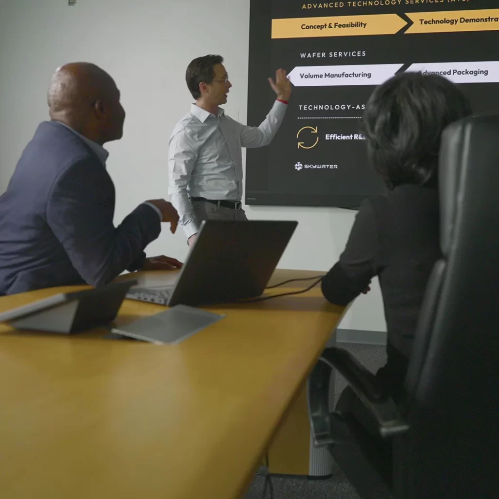 Three professionals in a conference room discuss a presentation slide about advanced technology services, with a focus on volume manufacturing and advanced packaging.