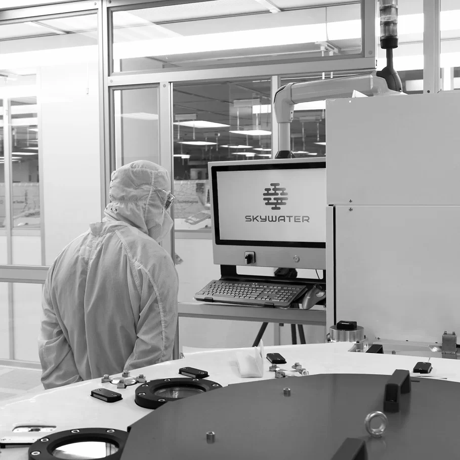 Worker in a cleanroom suit operating equipment in a SkyWater Technology semiconductor fabrication facility, with a computer screen displaying the SkyWater logo.